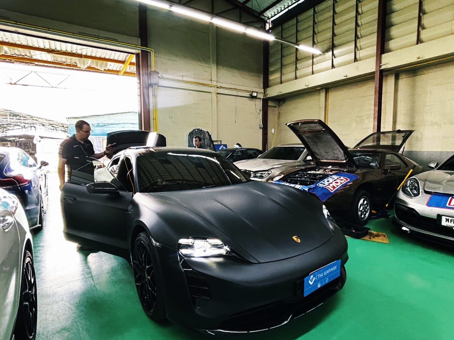 A Porsche Taycan in the CT14 Garage workshop, driver door open, a technician beside it with a tablet